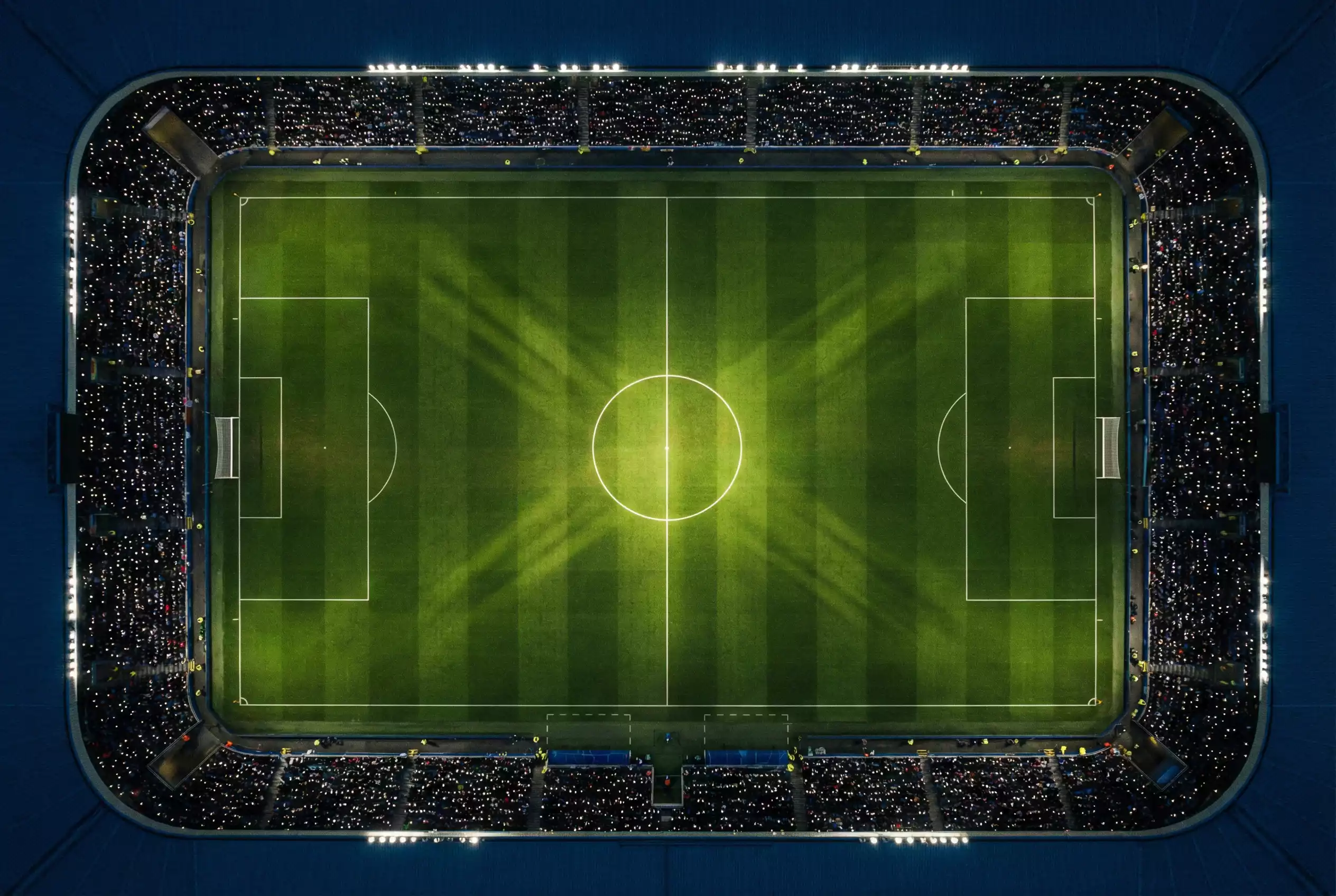 Campo de fútbol de noche iluminado por focos de estadio con el círculo central brillando sobre el césped